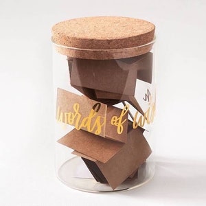 May include: A glass jar with a cork lid filled with small brown paper squares with gold lettering that reads "Words of Wisdom".