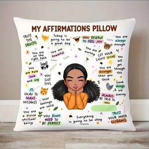 Puede incluir: Almohada blanca con un diseño colorido que presenta a una niña de dibujos animados y afirmaciones positivas. El texto incluye frases como "Confía en el proceso", "Eres poderoso", "Te mereces sentir alegría" y "Deja que tu luz brille".