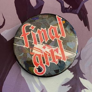 May include: A circular badge featuring the text "Final Girl" in red, decorative lettering. The badge has a shimmering, iridescent background. The background is purple with a raven perched on a branch.