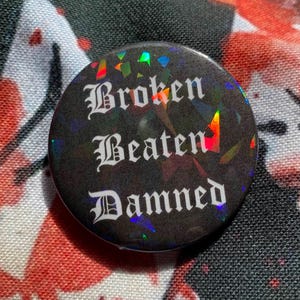 May include: A circular black button featuring the words "Broken Beaten Damned" in white gothic lettering. The button has a shimmering, iridescent surface. The background is a patterned fabric with red and black designs.