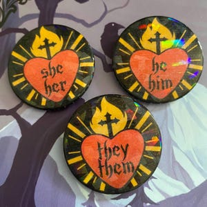 May include: Three circular badges with a black, iridescent finish. Each badge displays a red heart with a cross and flames, and the phrases "she her", "he him", and "they them" in gold lettering.
