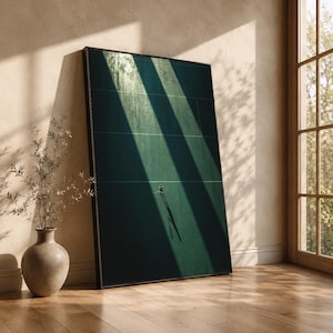 May include: A framed art print featuring a tennis court scene. The green court is bisected by strong shadows, with a silhouetted figure holding a racket. The artwork leans against a wall, next to a vase with decorative branches.