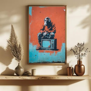 May include: Framed art print featuring a monkey sitting on a vintage television set. The artwork has a distressed, graffiti-style aesthetic with a red and blue color scheme. The monkey is wearing a red jacket and blue jeans.