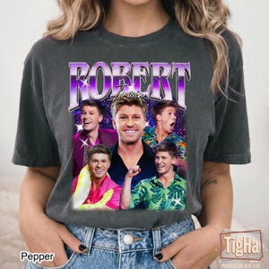 May include: A charcoal gray short-sleeve t-shirt featuring a graphic print of Robert Irwin. The design includes the name ROBERT in large purple letters, multiple photos of Robert Irwin, and a green leaf pattern. The shirt has a crew neckline.