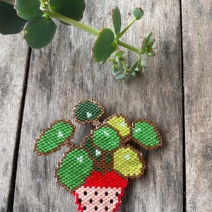 Op de afbeelding: Een kralenbroche in de vorm van een plant in een pot met ronde groene en gele bladeren, een rode pot met zwarte stippen en een metallic bronzen rand. Het kralenwerk heeft een rasterpatroon van kleine, uniforme kralen in groen, geel, rood en zwart.