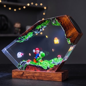 May include: A rectangular resin lamp featuring a wooden frame and a diorama of Mario riding Yoshi. The scene includes green moss, a white ghost, red-spotted mushrooms, floating gold coins, and a glowing yellow question block set within clear resin.