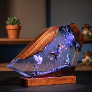 May include: A decorative lamp made of natural wood and clear resin, featuring an embedded miniature scene of a figure flying on a broomstick with a white owl and a castle, illuminated by internal purple and white LED lights on a wooden base.