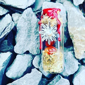Peut inclure: Une petite bouteille en verre avec un sceau de cire rouge et un charme de soleil argenté. La bouteille est remplie d'herbes séchées et d'une pierre rouge.