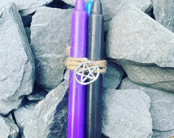 Può includere: Due candele, una viola e una nera, sono legate insieme con uno spago e un ciondolo a pentagramma d'argento. Le candele sono su un letto di pietre grigie.