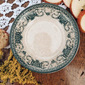 May include: A round ceramic plate featuring a cream-colored center and a wide border decorated with an intricate dark green floral and scrollwork pattern. The rim of the plate is finished with a fine, dotted texture along the outer edge.