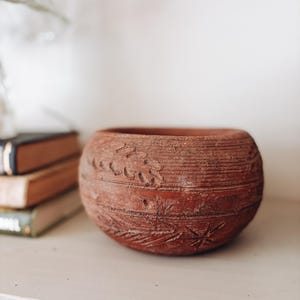 May include: A round, terracotta-colored clay bowl with a textured, matte surface. The exterior features horizontal grooved lines and carved, leaf-like botanical patterns encircling the body. The bowl has a wide, open top and a thick, rounded rim.