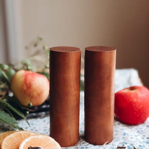May include: Two cylindrical salt and pepper shakers wrapped in smooth brown leather. Each shaker features a flat top with a series of small, circular holes arranged in a pattern for dispensing seasoning, standing upright on a flat surface.