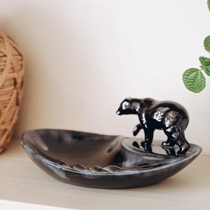 May include: A glossy black ceramic dish featuring a sculpted black bear figure standing on one side. The bowl has a smooth, curved interior surface and a dark, reflective finish, with the bear integrated into the rim of the shallow vessel.