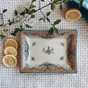 May include: Rectangular ceramic serving tray with a decorative border in orange, teal, and blue. The center features a blue and white illustration of a building. Lemons and greenery are arranged around the tray.