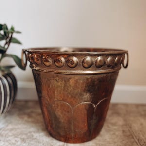 May include: A hammered copper bucket with a decorative rim featuring evenly spaced circular accents. The bucket has two small handles and a textured surface. The copper has a warm, aged patina, suggesting vintage or antique origins.