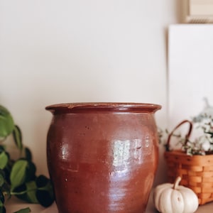 May include: A large, cylindrical brown ceramic crock with a smooth, glossy finish and a wide, slightly flared rim. The vessel features a deep, uniform earthy tone with subtle variations in the glaze, showcasing a sturdy, rounded base and a thick, solid construction.