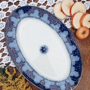 May include: An oval ceramic serving platter featuring a white center with a small blue floral motif and a wide, decorative border in shades of dark and light blue with a textured, sponge-like pattern and intricate white line detailing.