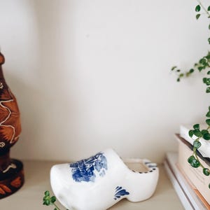 May include: A white ceramic clog with blue floral and landscape designs. The Dutch-style shoe is a decorative item, possibly a planter or trinket holder. The clog is on a neutral-colored surface, with a blurred background.