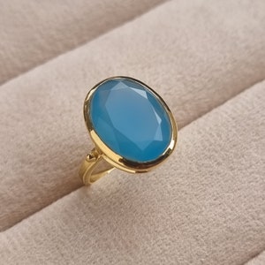 May include: A gold ring with a large oval blue gemstone. The ring is set in a gold band with a smooth finish.