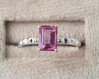 May include: A silver ring with a pink emerald-cut gemstone. The band is decorated with small triangular designs.
