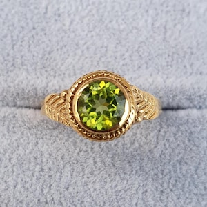 May include: A gold ring featuring a large, round, light green peridot gemstone. The ring has a detailed, textured gold band with a decorative design around the gemstone. The ring is set against a light gray background.