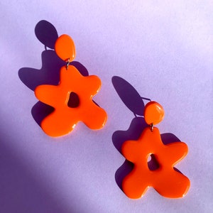 以下が含まれることがあります： 鮮やかなオレンジ色の花の形をしたイヤリングのペア。光沢のある仕上がりです。各イヤリングは、上部に小さな丸いスタッドがあり、下部に大きくて不規則な花の形をしています。薄紫色の背景に設定されています。