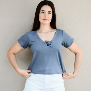 Puede incluir: Una mujer con una camisa azul de manga corta con un lazo en el escote y pantalones cortos de mezclilla blancos.