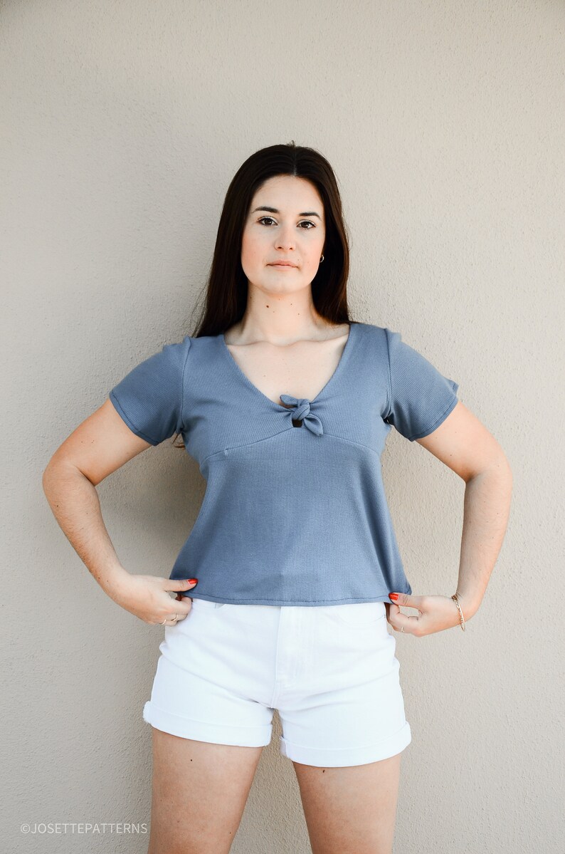 Puede incluir: Una mujer con una camisa azul de manga corta con un lazo en el escote y pantalones cortos de mezclilla blancos.