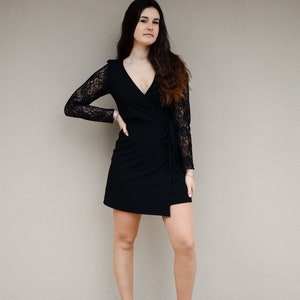 May include: A woman wearing a black wrap dress with lace sleeves and black ankle boots. She is standing in front of a beige wall.