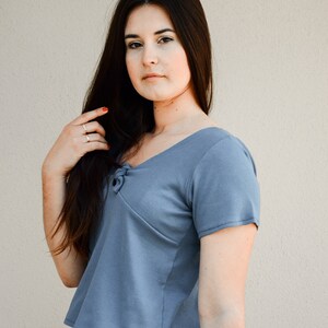 Puede incluir: Una mujer con un top azul corto de manga corta con un detalle anudado en el escote. Lleva pantalones blancos y tiene el pelo largo y oscuro.