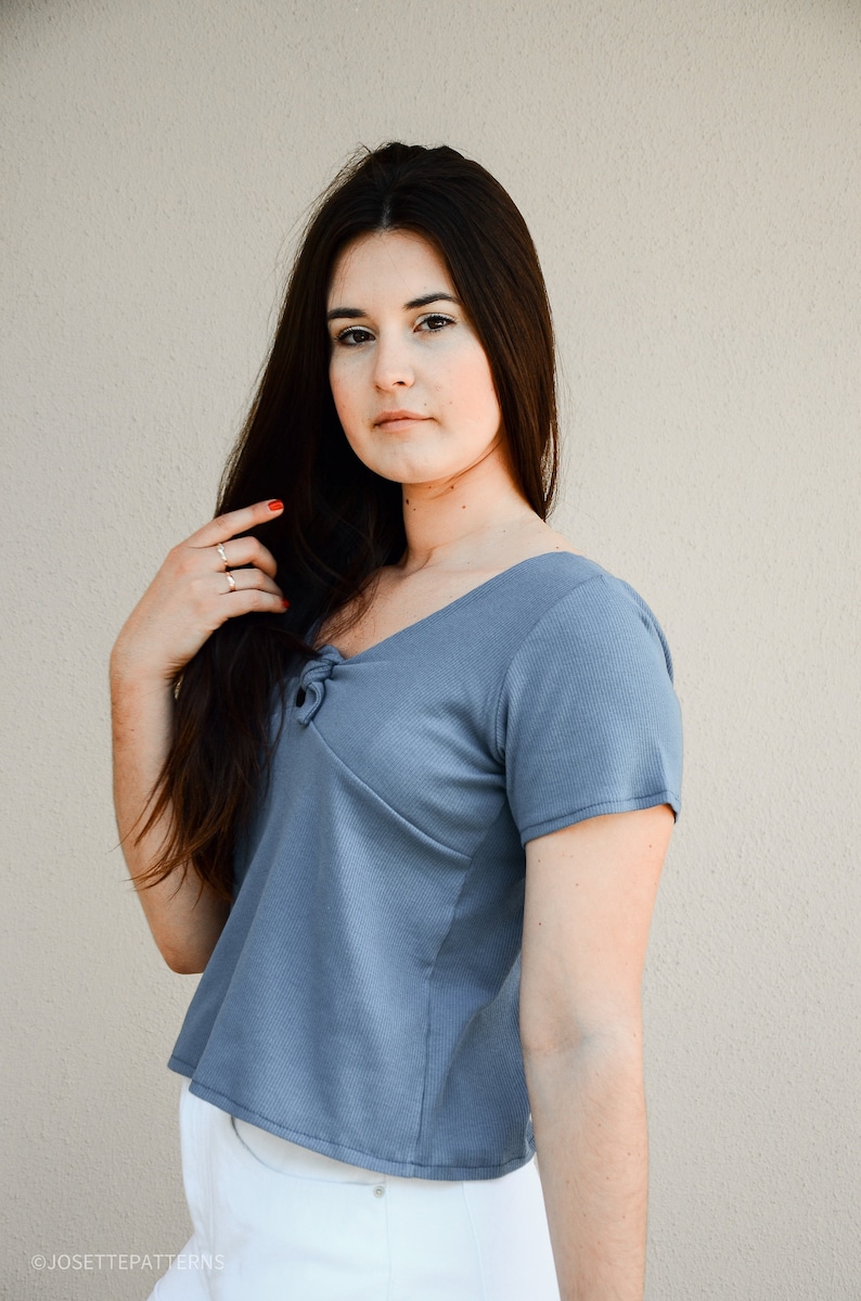 Puede incluir: Una mujer con un top azul corto de manga corta con un detalle anudado en el escote. Lleva pantalones blancos y tiene el pelo largo y oscuro.