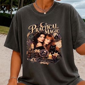 May include: A charcoal gray short-sleeve t-shirt featuring a graphic print with the text Practical Magic. The design includes two women, a black cat, a crystal ball, lit candles, and stacked books. The shirt has a crew neckline and a relaxed fit.