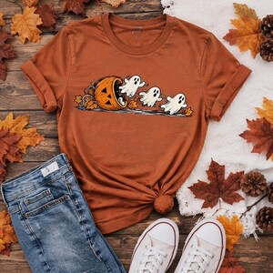 May include: A rust-colored short-sleeve t-shirt featuring a graphic print of a carved orange pumpkin with three white ghosts emerging from it, surrounded by scattered autumn leaves. The shirt has a crew neckline and rolled sleeves.