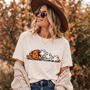 May include: A cream-colored short-sleeve t-shirt featuring a graphic print of three white ghosts emerging from a carved orange pumpkin surrounded by scattered autumn leaves. The shirt is paired with a beige knit cardigan and blue denim jeans.
