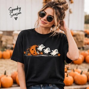 May include: A black short-sleeve t-shirt featuring a graphic of three white ghosts emerging from a tipped-over orange jack-o'-lantern surrounded by autumn leaves. The text "Spooky Season" with a heart icon is printed in the upper left corner.