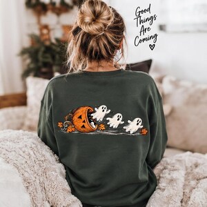 May include: A dark green long-sleeve sweatshirt featuring a graphic on the back of three white ghosts emerging from an orange jack-o'-lantern surrounded by autumn leaves. The text "Good Things Are Coming" with a heart is printed above.