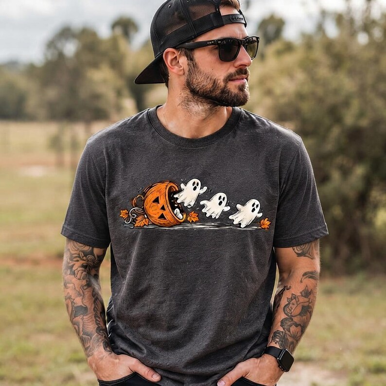 May include: A charcoal gray short-sleeve t-shirt featuring a graphic print of three white ghosts emerging from a carved orange jack-o'-lantern pumpkin surrounded by small autumn leaves on the ground, centered on the chest of the garment.