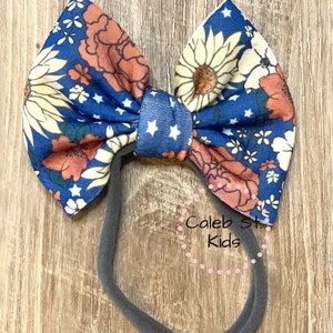 Pode incluir: Uma faixa de cabelo com estampa floral azul e laranja com estrelas brancas. A faixa de cabelo &eacute; amarrada em um la&ccedil;o e tem uma banda el&aacute;stica preta.