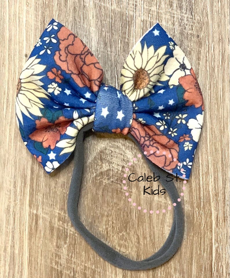 Pode incluir: Uma faixa de cabelo com estampa floral azul e laranja com estrelas brancas. A faixa de cabelo &eacute; amarrada em um la&ccedil;o e tem uma banda el&aacute;stica preta.
