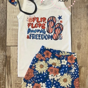 Pode incluir: Um top branco sem mangas com um gr&aacute;fico vermelho, branco e azul que diz "Flip Flops, Fireworks & Freedom". O top &eacute; combinado com shorts azuis com estampa floral e estrelas brancas. Inclui uma faixa de cabelo com estampa de girassol.