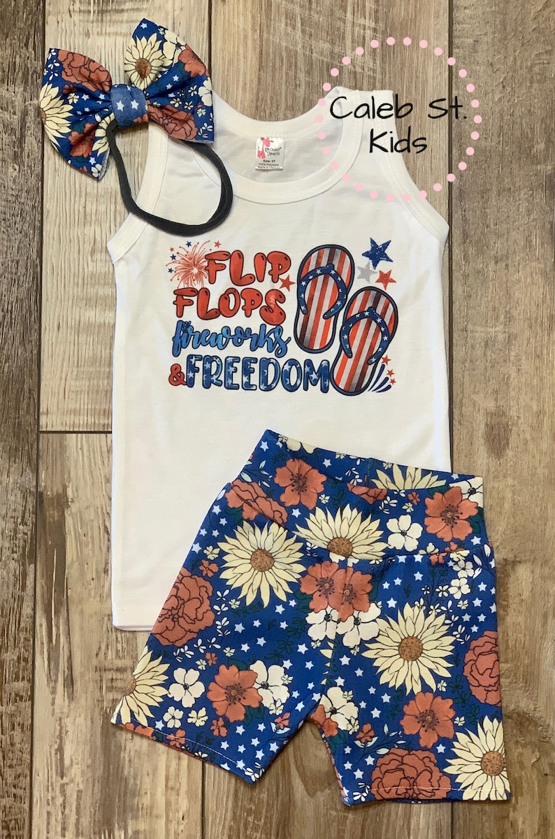 Pode incluir: Um top branco sem mangas com um gr&aacute;fico vermelho, branco e azul que diz "Flip Flops, Fireworks & Freedom". O top &eacute; combinado com shorts azuis com estampa floral e estrelas brancas. Inclui uma faixa de cabelo com estampa de girassol.