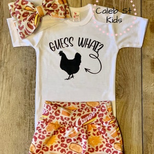 May include: A white baby onesie with the text "Guess What?" and a black silhouette of a chicken with a tail feather pointing to the right. The onesie is paired with a leopard print diaper cover with the text "Canes" printed on it. The onesie and diaper cover are on a wooden surface.