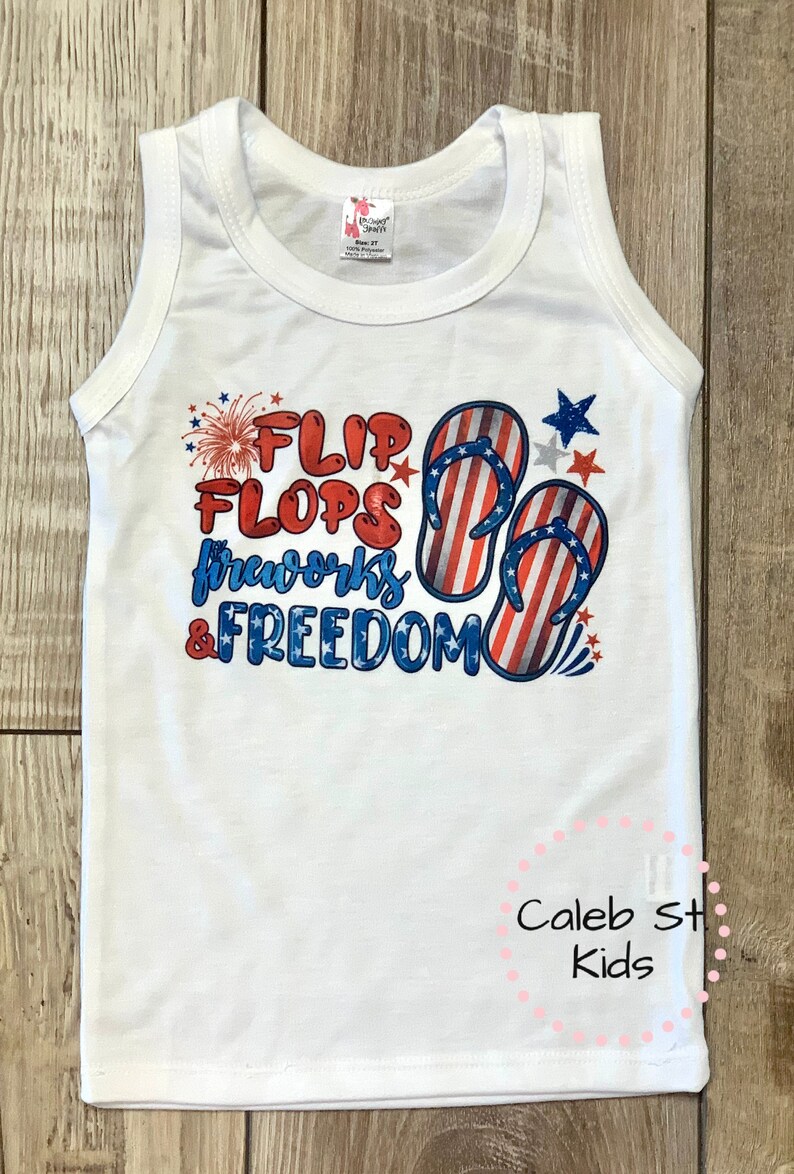 Pode incluir: Camisola branca sem mangas com um gr&aacute;fico vermelho, branco e azul que diz "Flip Flops, Fireworks & Freedom". O gr&aacute;fico inclui um par de chinelos com um design de bandeira americana e fogos de artif&iacute;cio.