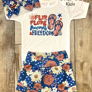 Pode incluir: Um macac&atilde;o branco para beb&ecirc; com o texto "Flip Flops Fireworks & Freedom" e um gr&aacute;fico de chinelos vermelhos, brancos e azuis. Tamb&eacute;m est&aacute; inclu&iacute;do uma faixa de cabe&ccedil;a de estampa floral azul e branca e shorts de estampa floral azul e branca.
