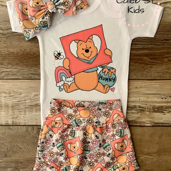 May include: A white baby onesie with a Winnie the Pooh graphic. The onesie has a pink heart with a picture of Winnie the Pooh inside. The text "Hunny" is on the onesie. A matching pair of shorts with a Winnie the Pooh print. The shorts have a pink, yellow, and green color scheme. The shorts have a white elastic waistband. The onesie and shorts are part of a baby outfit.