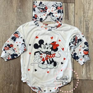 May include: White baby bodysuit and matching headband set featuring Mickey and Minnie Mouse. The bodysuit has long sleeves with a Mickey and Minnie pattern, and a large graphic of the couple. Red hearts and Mickey Mouse silhouettes are scattered across the design.