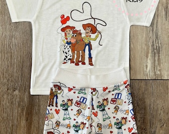 May include: White t-shirt with a graphic of Woody, Jessie, and Bullseye from Toy Story. Matching white shorts with a repeating pattern of Toy Story characters, hearts, and the words "XOXO" and "U".