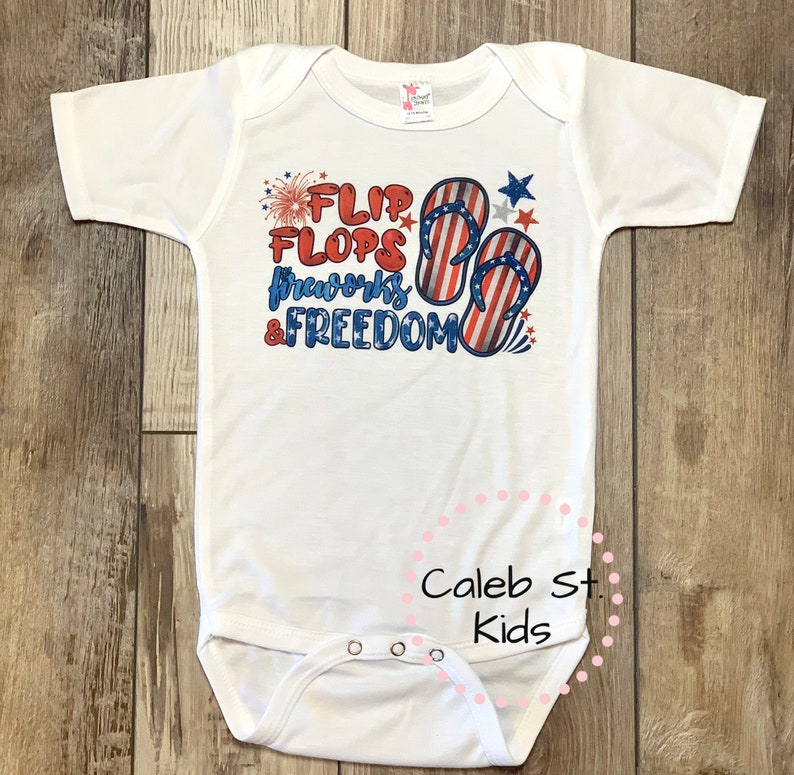 Pode incluir: Um macac&atilde;o branco para beb&ecirc; com um gr&aacute;fico vermelho, branco e azul que diz "Flip Flops, Fireworks & Freedom". O gr&aacute;fico inclui um par de chinelos com um design de bandeira americana e fogos de artif&iacute;cio. O macac&atilde;o tem uma etiqueta rosa que diz "Caleb St. Kids".