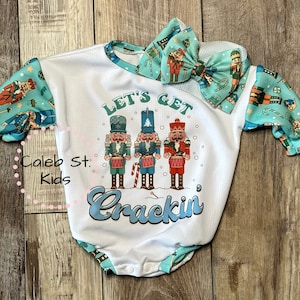 May include: White baby onesie with a blue and green nutcracker print on the sleeves. The onesie has the text "Let's Get Crackin'" with three nutcracker figures playing drums.