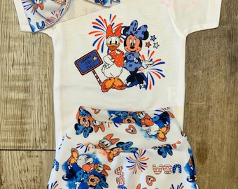 May include: A white, short-sleeved shirt with a red, white, and blue print of Mickey Mouse and Daisy Duck celebrating the 4th of July. The shirt is paired with white shorts with a red, white, and blue print of Mickey Mouse and Daisy Duck. A white headband with a red, white, and blue bow is included.